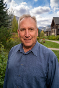 Portrait of Ross Jansen of Spring Creek Real Estate in Canmore, wearing a blue collared shirt, and standing and smiling in front of green foliage with mountains in the background.